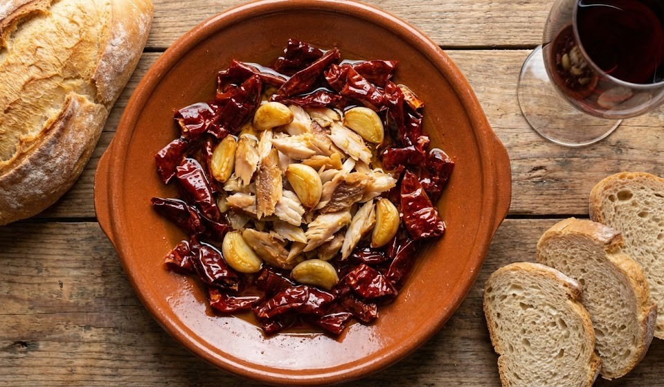 Plato de barro rústico con Pericana Alcoyana tradicional, pimientos secos fritos, bacalao desmigado y ajos en aceite de oliva, sobre mesa de madera con pan y vino